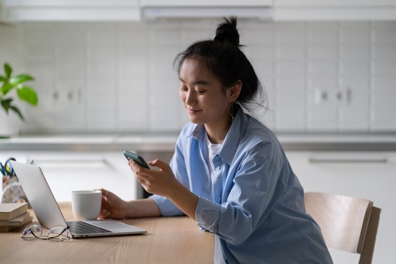 A woman sitting at a desk in front of her laptop, holding her phone 