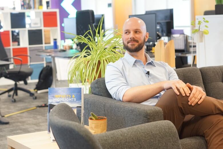 A bearded man sits on a couch with empty desks behind him. 