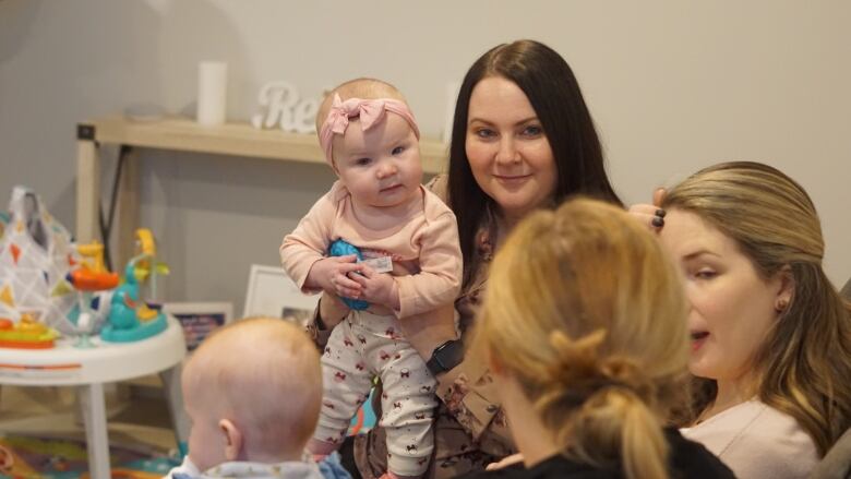 Mothers sit together, talking, holding babies. 