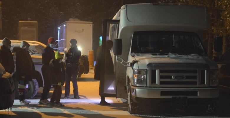 Adults and children stand in line to board a bus at night in a snowy parking lot.