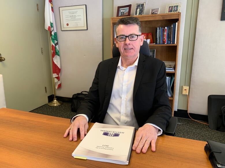 A man in a suit jacket sits at a desk in an office with a report on the desk in front of him.