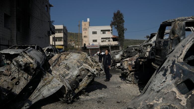 Two men surrounded by burned out cars use their phones to take photos of wreckage. 