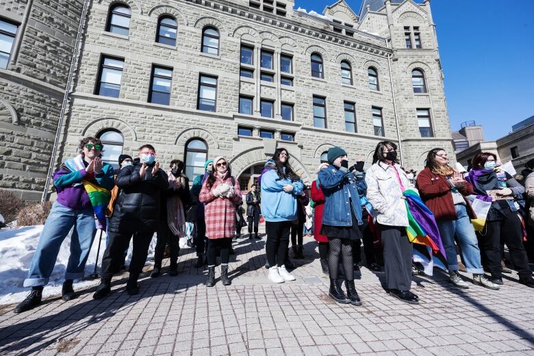 A group of people, some holding rainbow or transgender-pride flags, stand side by side at a rally.