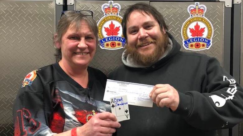 A woman and man pose for a photo, the woman holding up an ace of clubs with Brad Sanderson written on it with blue marker. The man holds up a cheque for $16,368.