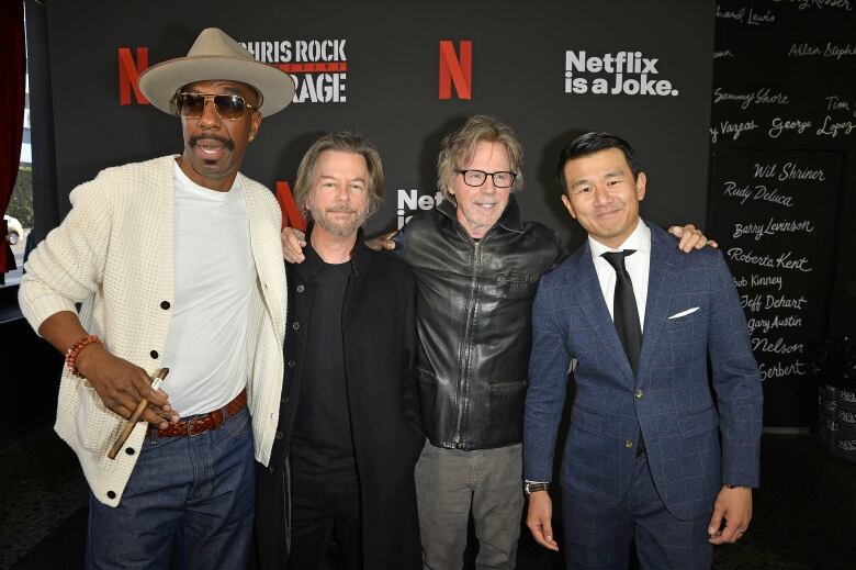 Four men stand shoulder-to-shoulder as they pose for a photo before a backdrop that says, 