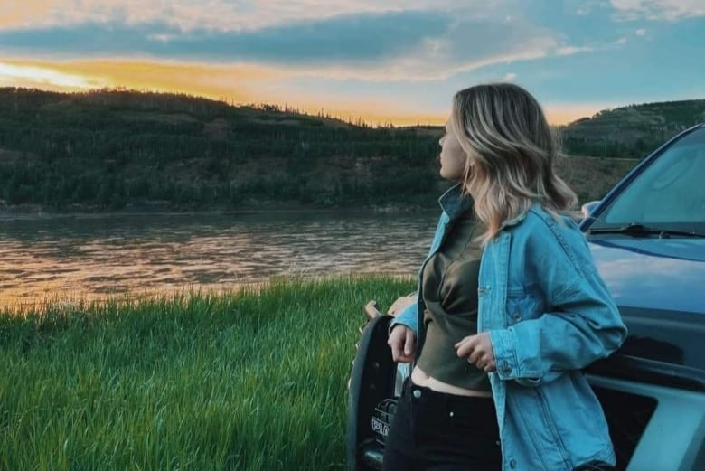 A blonde woman leans against a car while standing next to a river during a sunset.