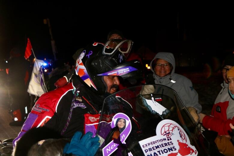 A man wearing red and purple gear drives a Ski-Doo. People greet him with smiles.