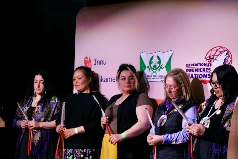 Five women stand on a stage holding feathers 