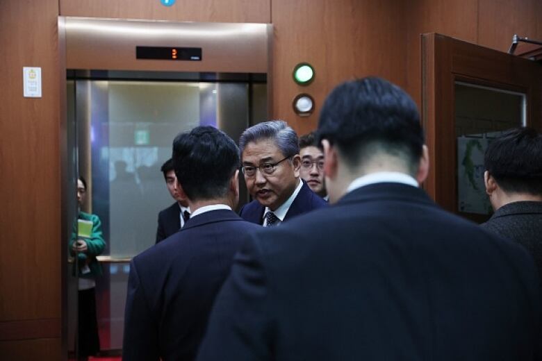 A man facing the camera, with grey hair and glasses, talks with other men in suits, their backs to the camera, in front of an elevator.