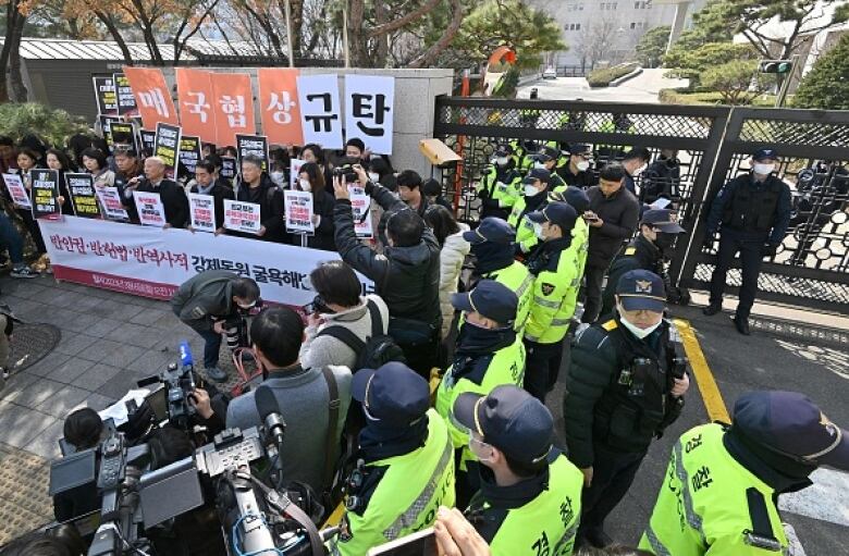 Photographers shoot a group of protesters carrying signs. A row of police stand behind the photographers.