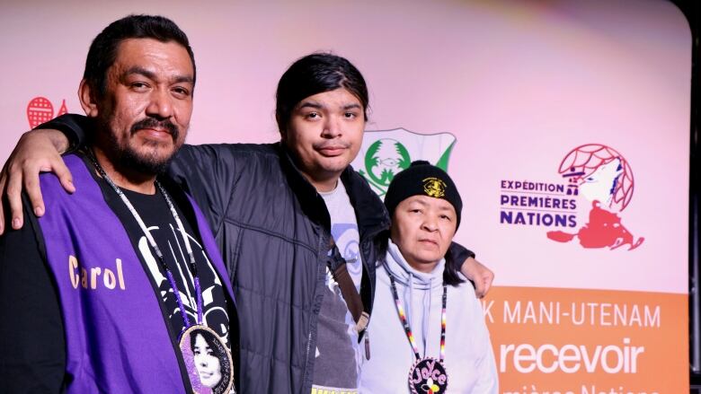 Three people stand next to eachother looking at the camera. One of them is wearing a purple vest. They stand in front of a sign that says First Nations Expedition in French.