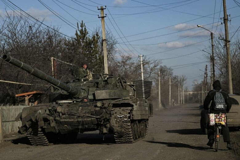 A tank with a soldier atop it is shown travelling on a road as a bicyclist heads in the opposite direction.