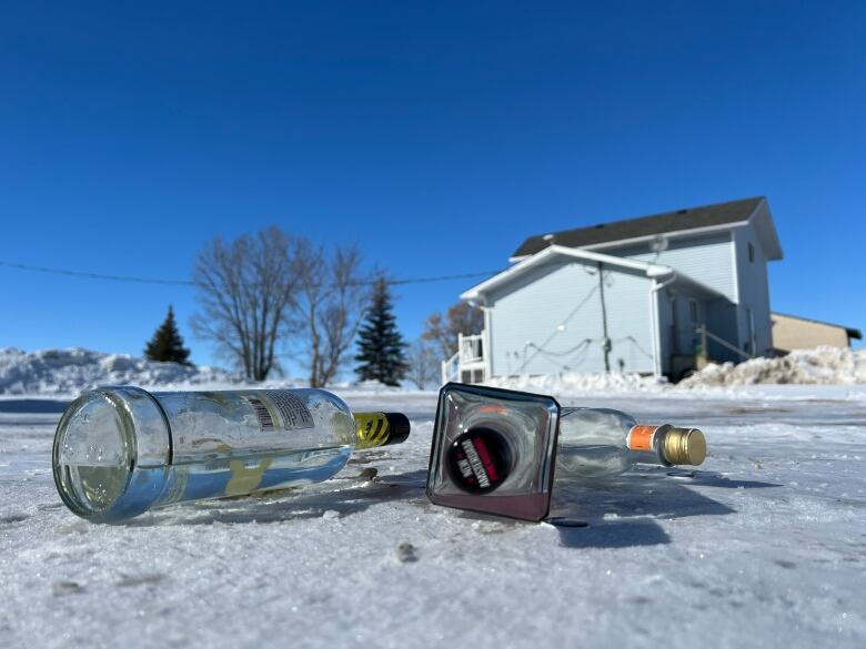 Three alcohol bottles are seen on a snowy ground with a house and trees in the background.