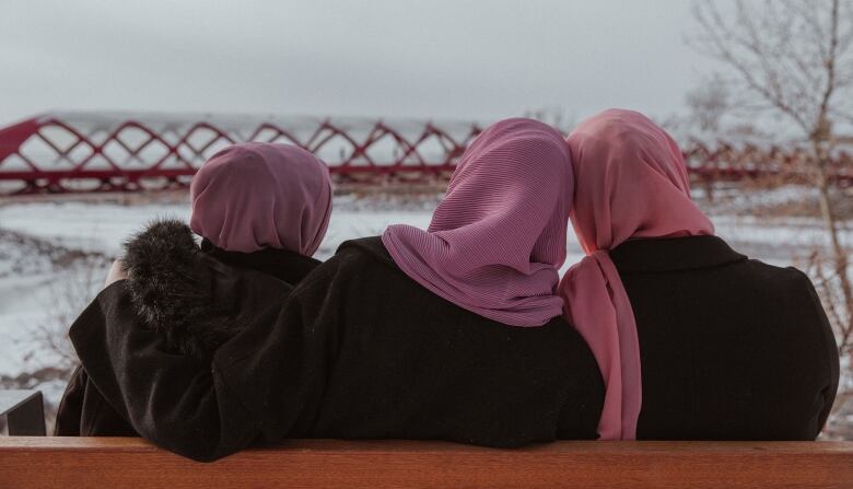 The back of three women's heads, all wearing different textured and coloured hijabs, with Calgary's Peace Bridge in the background