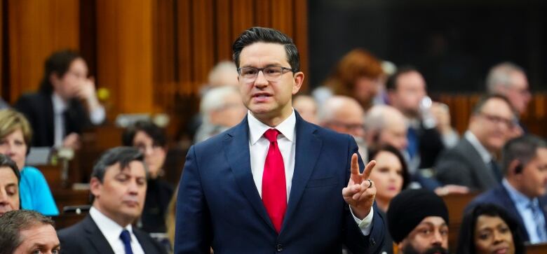 A man in a suit stands and gestures while addressing a large room of people.