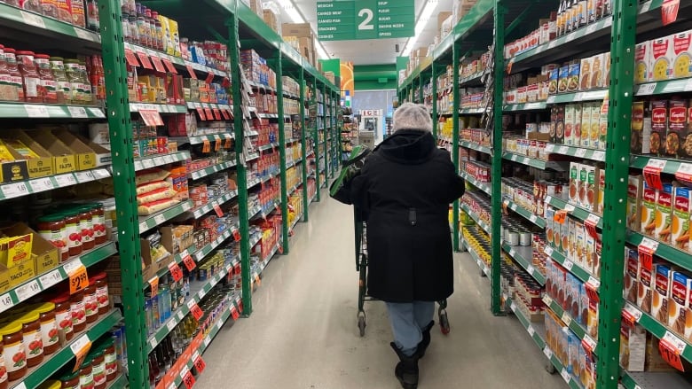 A woman wearing winter clothes pushes a cart down a grocery store aisle 