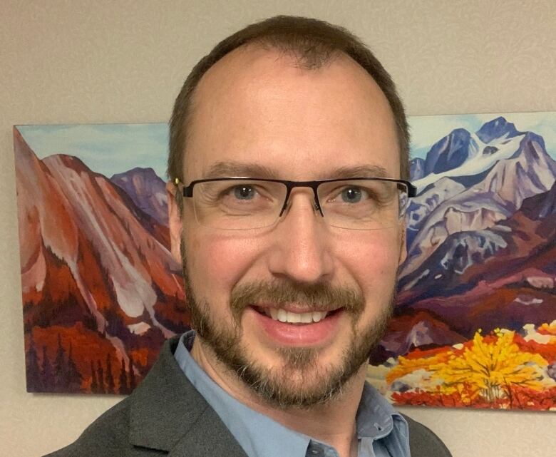 A man wearing glasses is smiling directly at the camera, standing in front of a painting. 