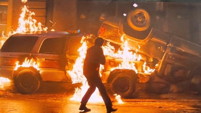 A man stands in front of a police car and pickup truck which are on fire after a collision and explosion. 