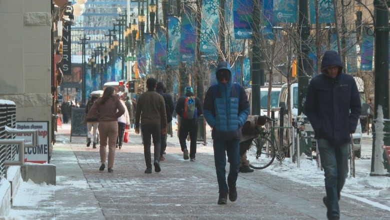 People wearing jackets walk along a snowy sidewalk with their heads down and their hands tucked into their pockets. 