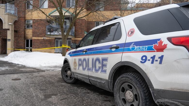 A police car sits parked by an apartment complex with snow on the ground and yellow police tape wrapped around a tree.