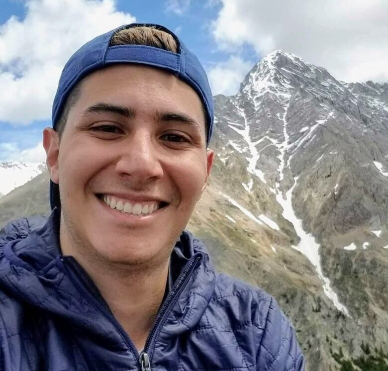 A guy in a dark blue jacket and backwards cap smiles at the camera with a mountain behind him.