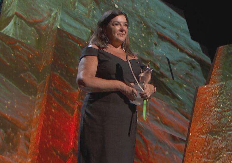 A woman wearing a dress holds onto a trophy and eagle feather while on a well-lit stage. 