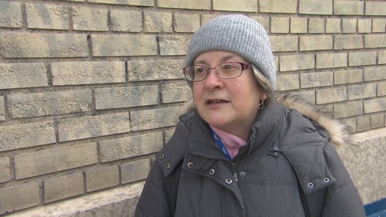 A woman wearing a coat with a fur-lined hood, a toque, and glasses is pictured standing next to a brick building.