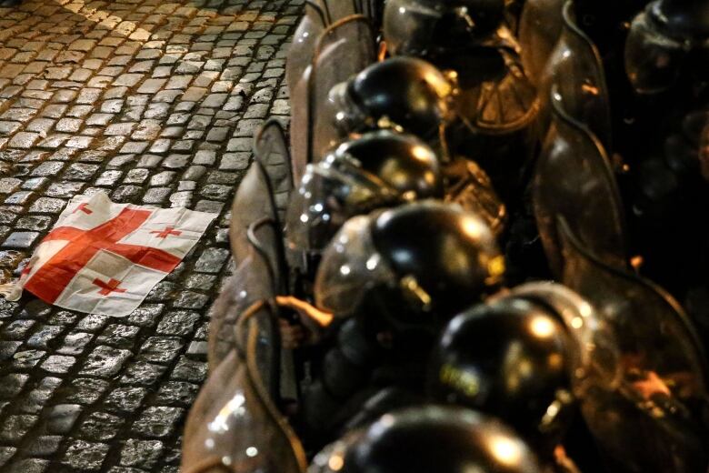 A line of helmeted and shielded riot police stands with the Georgian flag - a large red cross on a white background, with four smaller red crosses in each of the quadrants - lying on the cobblestones in front of them.