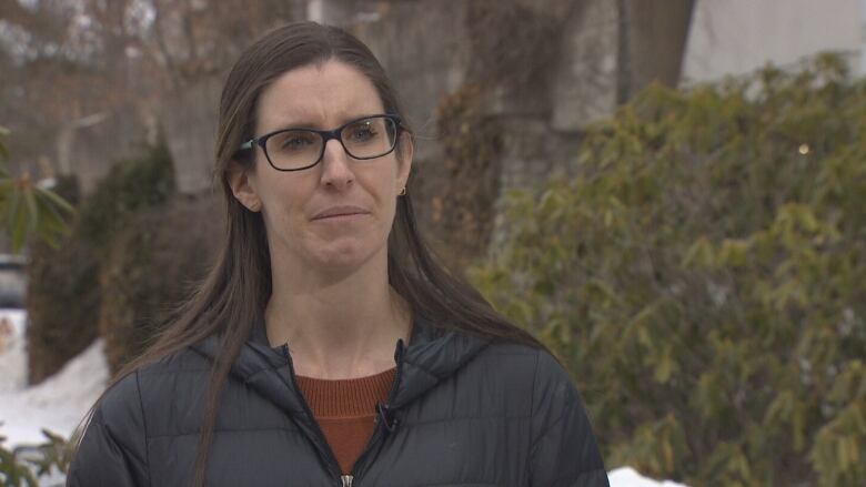 A woman with long brown hair and square glasses looks at the camera frowning. She is outdoors, in front of a tree, and wearing a black puffy jacket.