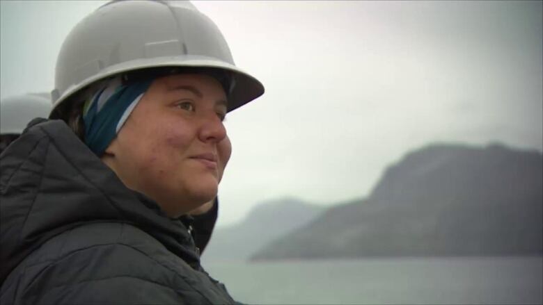 A woman wearing a hard hat and a puffer jacket looks over the ocean with mountainous slopes peeking through the background.