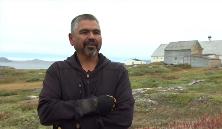 A man in a hooded windbreaker is pictured in front of a green expanse, punctured by three buildings, one visibly battered, a long white building, and a small cabin. 