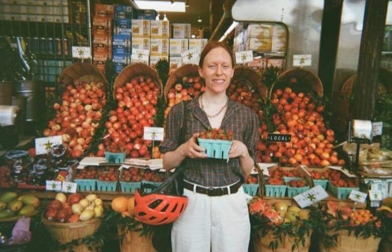 A person smiles in front of fresh fruit, while holding a basket of strawberries.