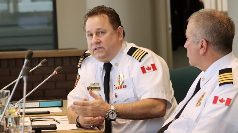 Two men in white firefighter uniforms speak in front of microphones.