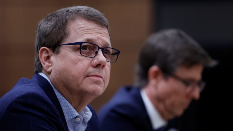 A man with brown hair, glasses and wearing a suit sits at a small microphone, next to another man in the background, and looks up.