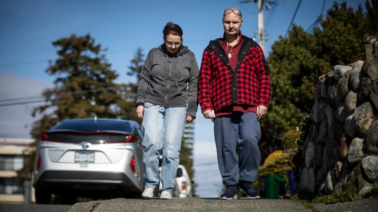 A woman and a man walk down a sidewalk together on a sunny day. Both are dressed for cool weather; the woman is looking down at her feet as she walks.