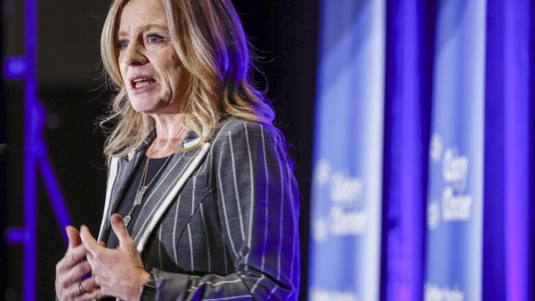 Alberta NDP Leader Rachel Notley interlaces her fingers as she speaks at a Calgary event, in purple light and in front of Calgary Chamber of Commerce banners.