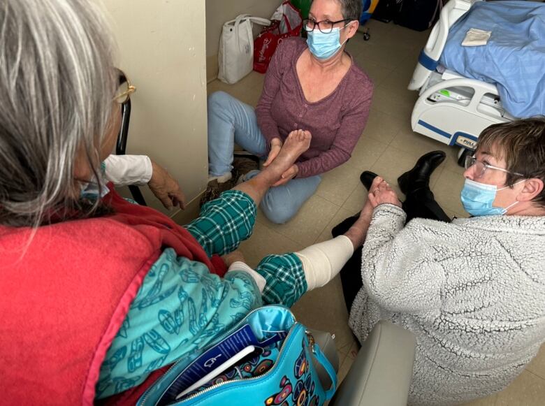 Two women sit on the floor, rubbing the feet of an older woman.