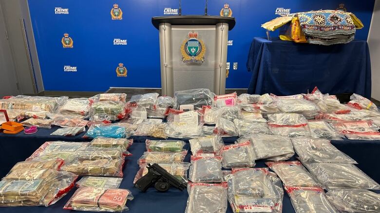 Bags of cash and drugs are seen on a table in front of a podium in the centre of the frame. Off to the left is a stack of rugs. In the background is a Winnipeg police banner.