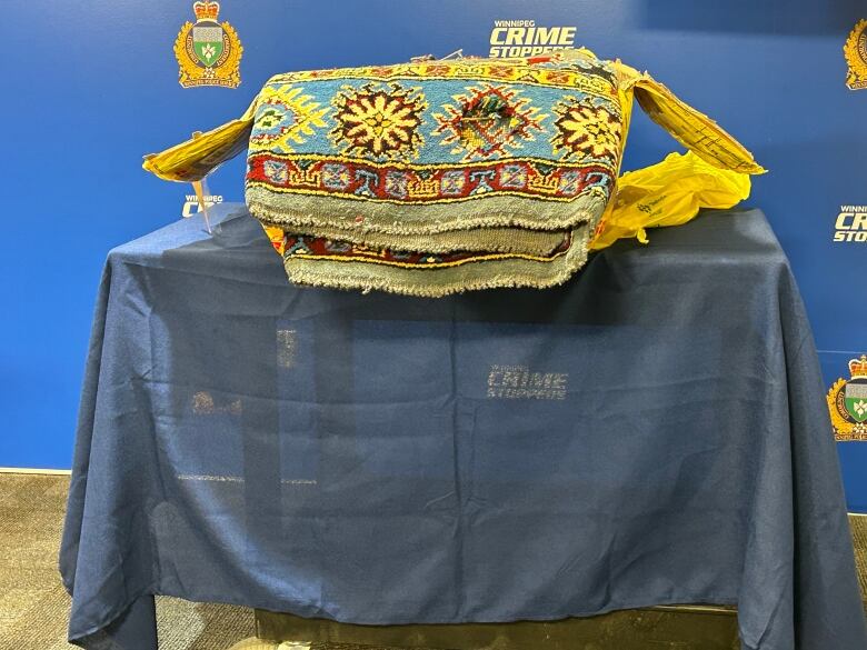 A stack of intricately woven blue and yellow rugs stand on a table with a faded 