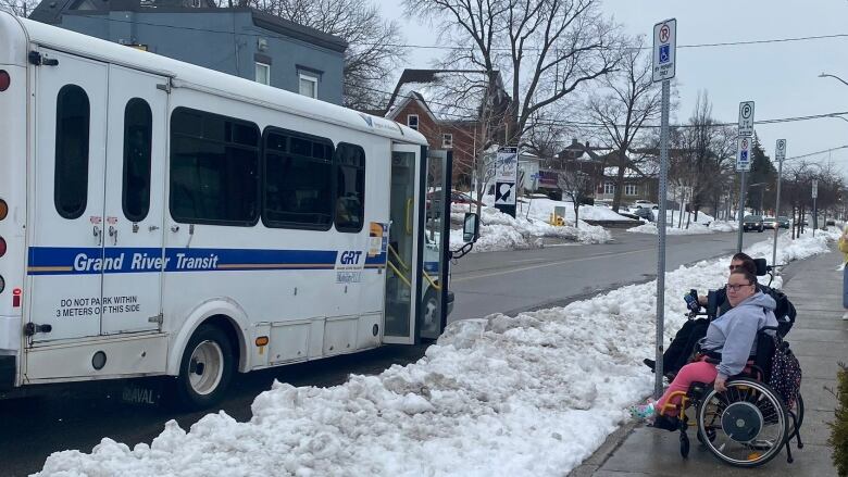 People in wheelchairs on sidewalk and a mobility bus on road, but a large snowbank is between them and the people can't get on the bus.