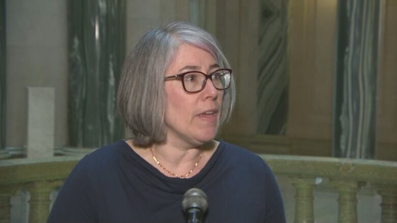 A woman with grey hair and glasses stands in front of a microphone at the rotunda of the legislature building in Regina, Sask. 