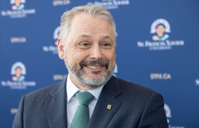 A man with short hair and a beard wearing a suit stands in front of a blue banner.