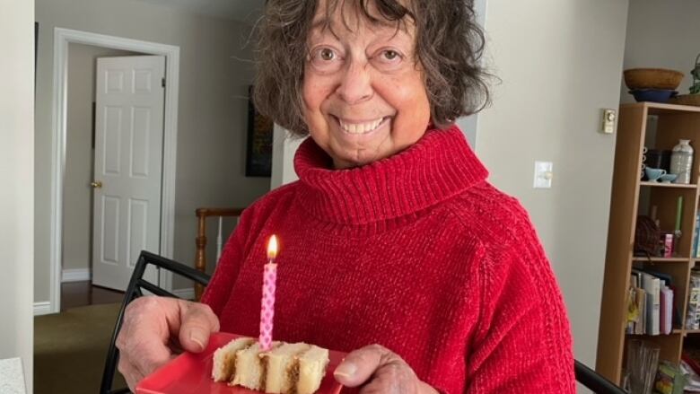 A woman with short, dark and wavy hair smiles at the camera while holding a square red plate containing a cake with a single pink candle that is lit. She is smiling at the camera and wearing a red knit turtleneck sweater.