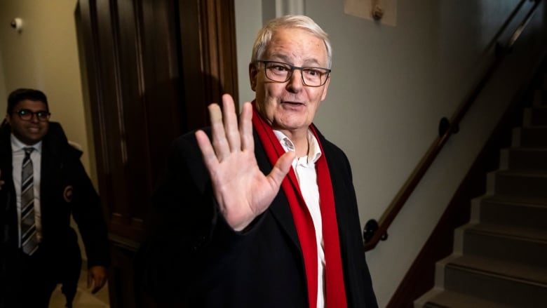 A man wearing a coat and a red scarf waves to the camera.