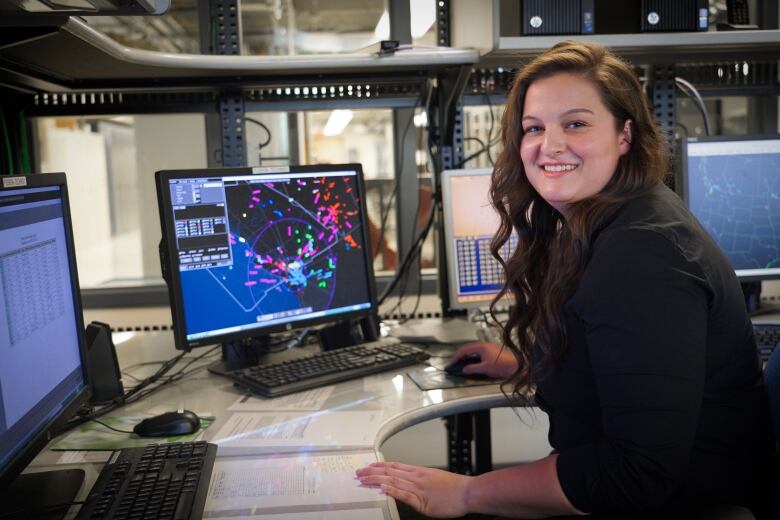 A woman smiles at the camera while the computer screens in front of her show the flight paths of aircraft.