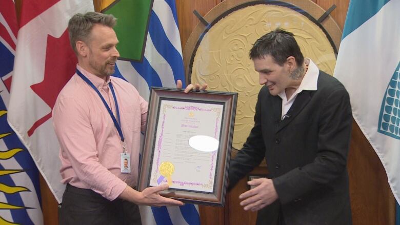 A man hands another man a framed official proclamation text.