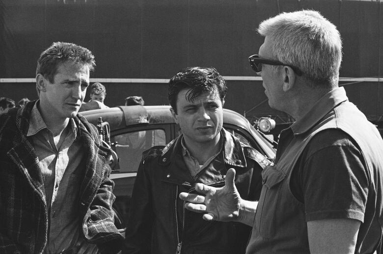 Two men are shown listening while an older man in sunglasses speaks and gestures, ina black and white photograph.