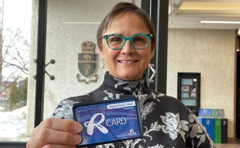 A woman in a black and white turtleneck sweater, wearing green glasses, holds up a blue card with R-Card written on it. 