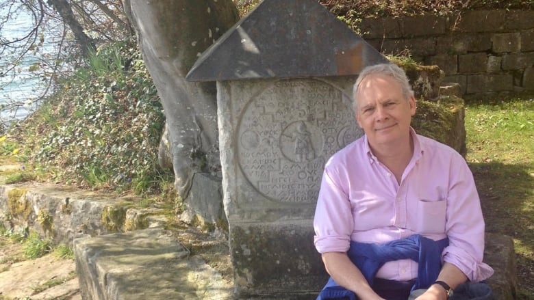 A man with white hair and wearing a pink shirt stands outside in front of a stone tablet.