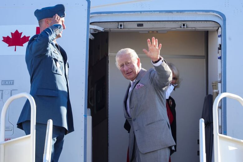 A person waves from the top of the steps up to the entrance of plane, with a person saluting standing in front of them.